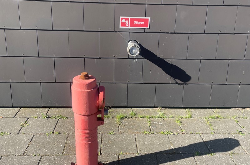 Billede af en brandhane som står på en flisebelagt overflade, lige bagved er der en side af et hus hvor der er udgang til vand hvor der står stigrør.